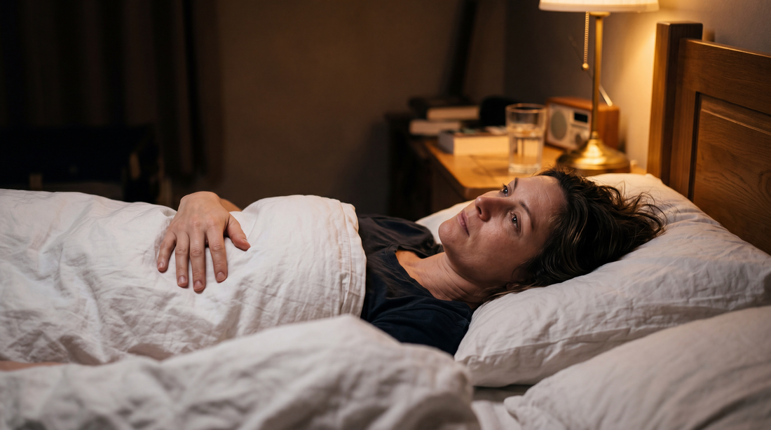 Woman in her forties lying in bed at night with one hand resting on her stomach, soft bedside lamp light